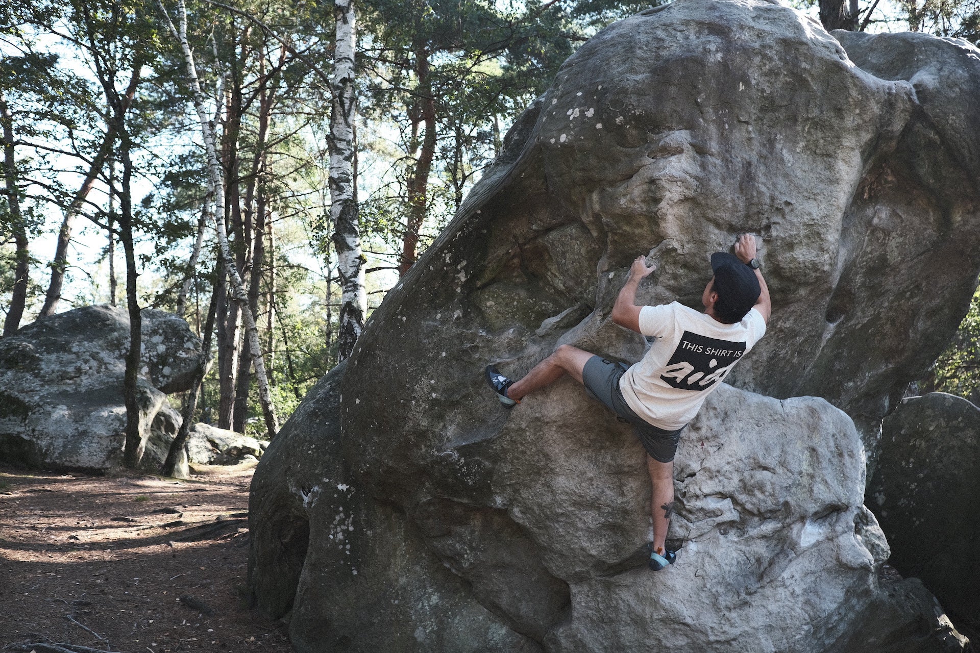 Person climbing a boulder in the woods while wearing a shirt that says "this shirt is aid"