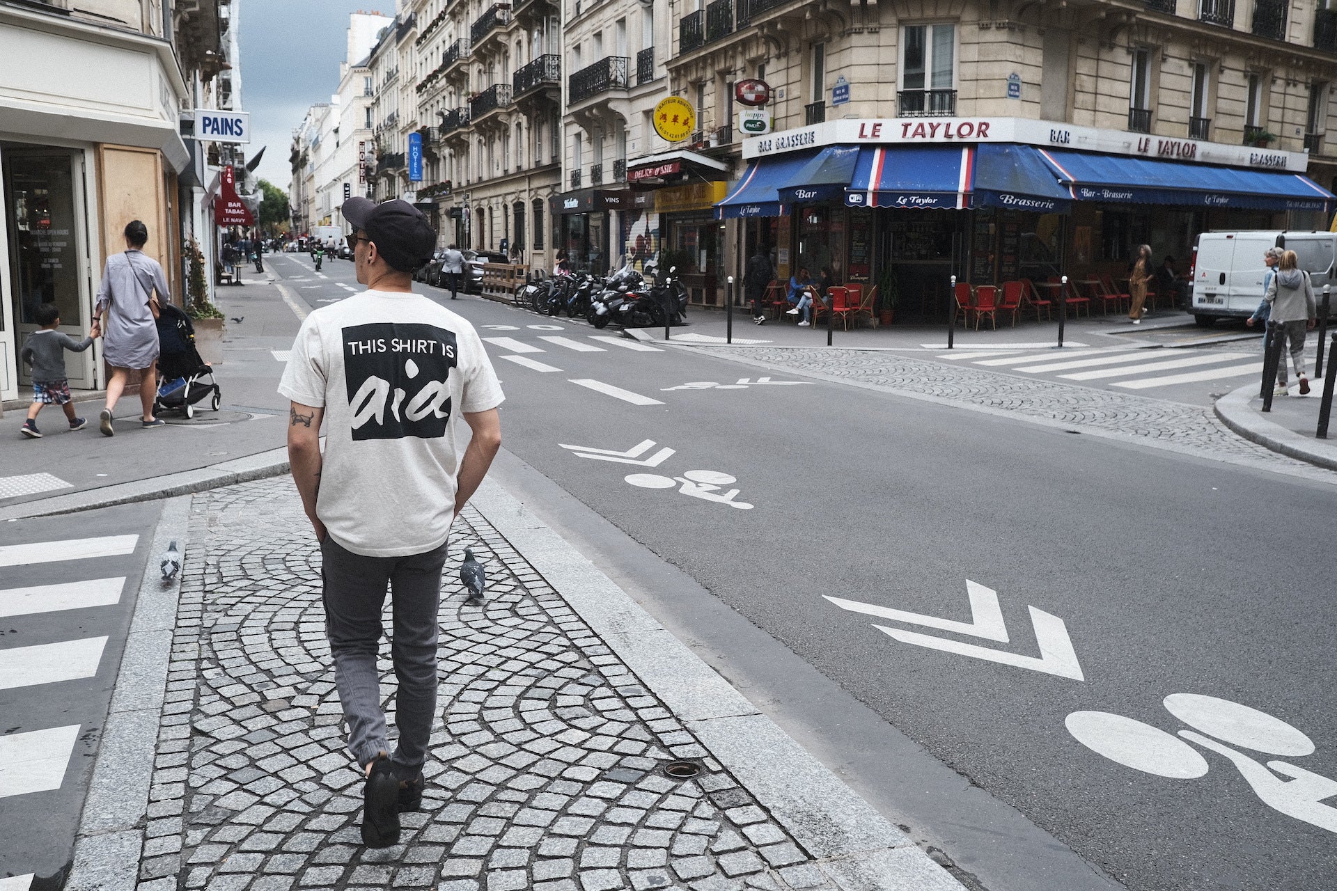 A person walking on an urban street, wearing a shirt that says "this shirt is aid" on the back