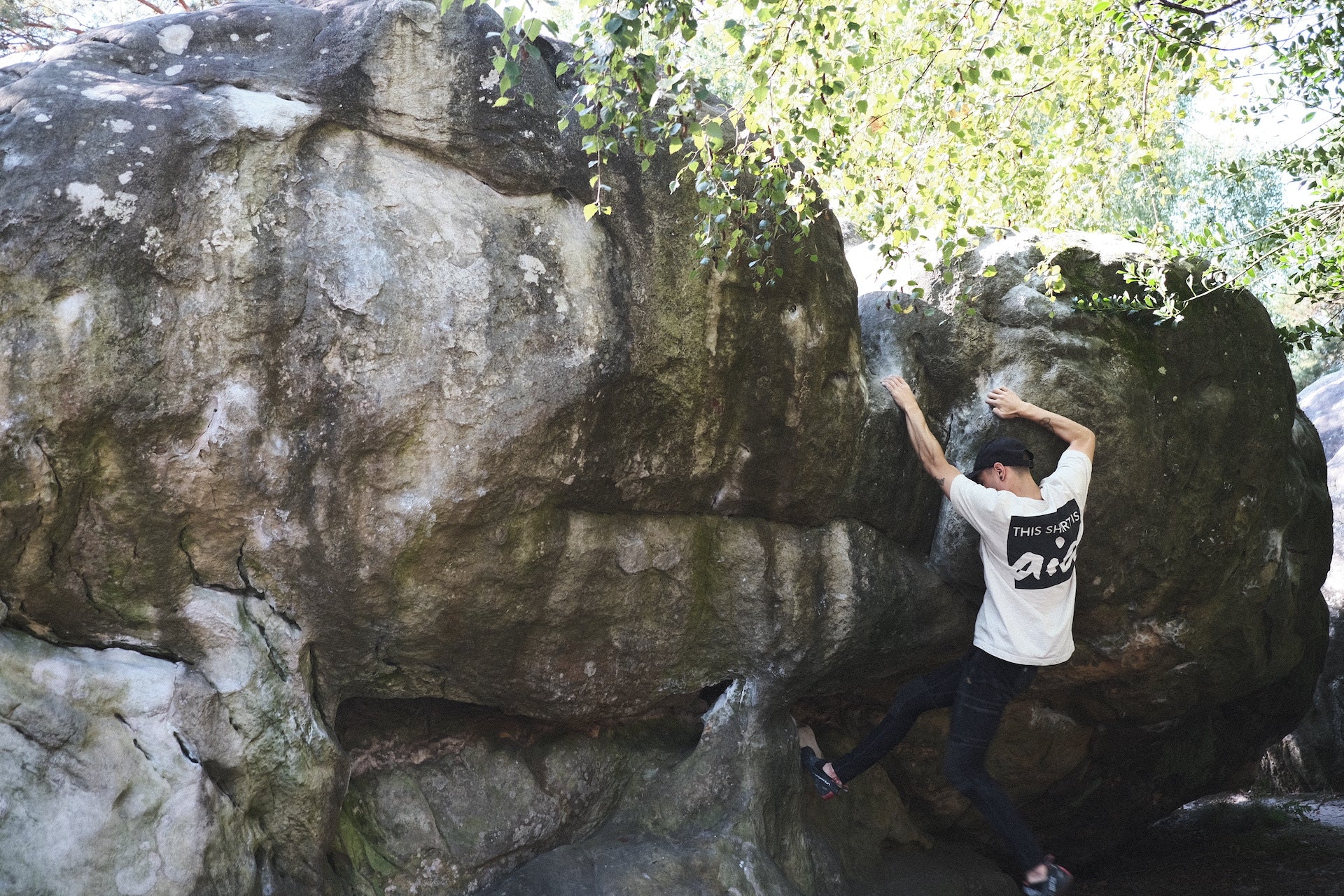 A person climbing a boulder in the woods, wearing a shirt that says "this shirt is aid" on the back